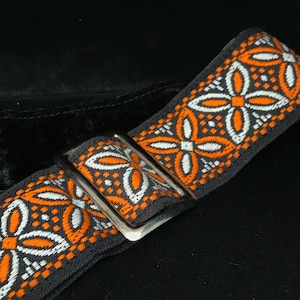 May include: A dark gray fabric strap featuring a repeating embroidered pattern of orange and white four-petaled floral shapes. The strap includes a rectangular silver-toned metal slider buckle used for adjusting the length of the strap.