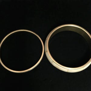 Puede incluir: Dos brazaletes dorados. Uno es una banda simple, el otro presenta una incrustaci&oacute;n de color crema. Los brazaletes se exhiben sobre un fondo negro, resaltando su brillo met&aacute;lico y colores contrastantes. Estos accesorios son adecuados para el uso diario.