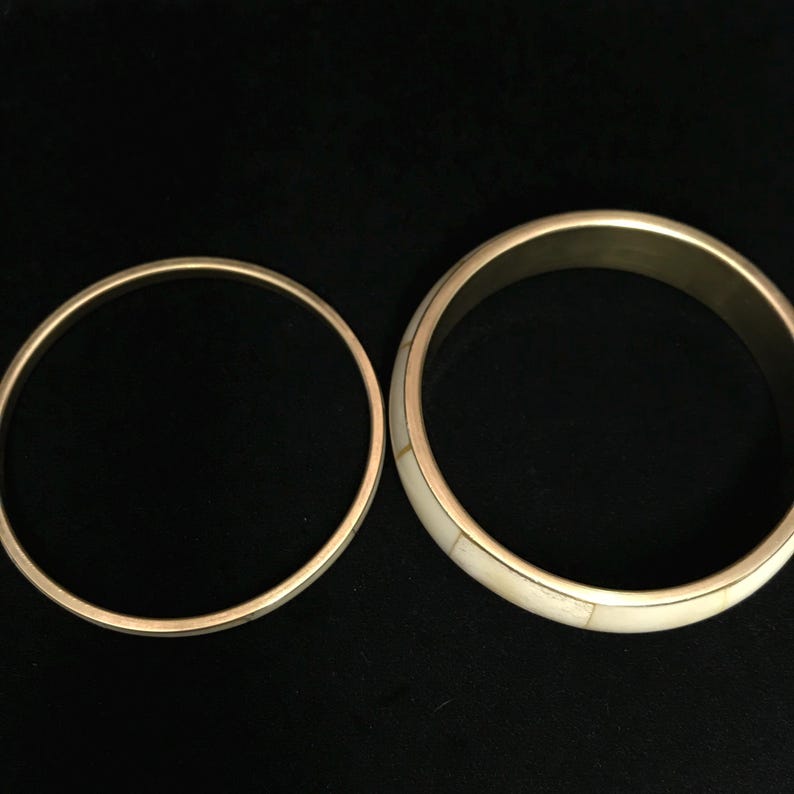 Puede incluir: Dos brazaletes dorados. Uno es una banda simple, el otro presenta una incrustaci&oacute;n de color crema. Los brazaletes se exhiben sobre un fondo negro, resaltando su brillo met&aacute;lico y colores contrastantes. Estos accesorios son adecuados para el uso diario.