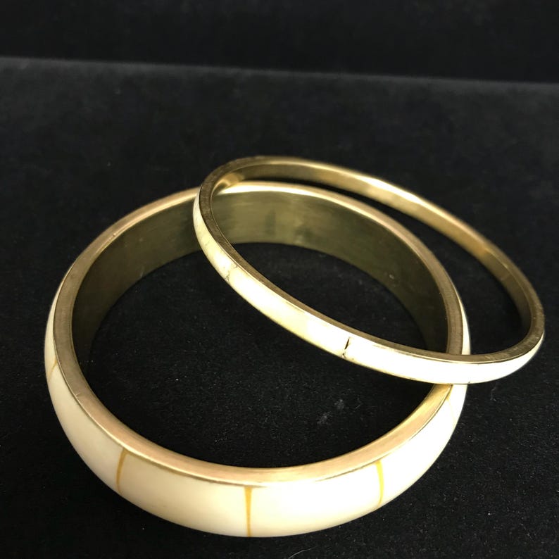Puede incluir: Dos brazaletes dorados con incrustaciones de color marfil y finas rayas amarillas. Un brazalete es m&aacute;s ancho que el otro. Los brazaletes est&aacute;n colocados sobre un fondo oscuro.