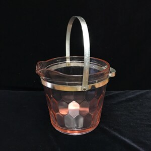 May include: A pale pink glass ice bucket featuring a textured honeycomb pattern on the exterior. The bucket is encircled by a polished silver-tone metal band and includes a matching silver-tone metal swing handle attached to the sides.