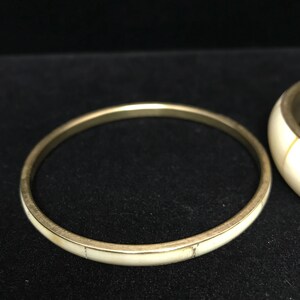 Puede incluir: Una pulsera de brazalete dorada con una incrustaci&oacute;n de color crema. La pulsera es circular y parece estar hecha de metal. La incrustaci&oacute;n de color crema tiene un aspecto veteado. La pulsera se muestra sobre una superficie negra.