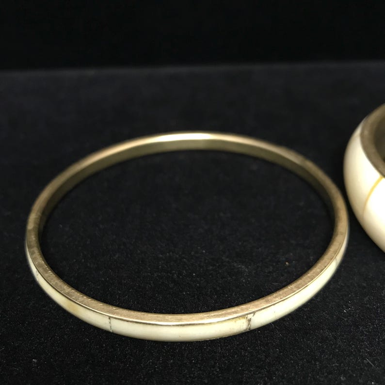 Puede incluir: Una pulsera de brazalete dorada con una incrustaci&oacute;n de color crema. La pulsera es circular y parece estar hecha de metal. La incrustaci&oacute;n de color crema tiene un aspecto veteado. La pulsera se muestra sobre una superficie negra.