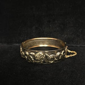 May include: A gold-toned hinged bangle bracelet with an ornate floral and grape design. The bracelet has a detailed pattern of leaves, flowers, and grape clusters. A small gold chain is attached to the clasp.