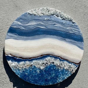 May include: A circular decorative piece featuring a fluid art design with swirling layers of navy blue, light blue, and white. The surface is accented with textured, crushed glass elements in clear and blue tones along the top and bottom edges.
