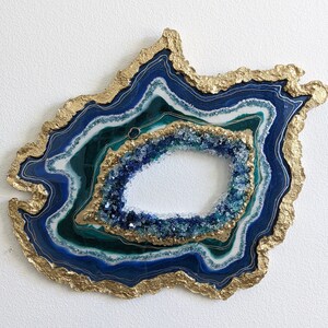 May include: A decorative geode slice with a gold-colored, textured edge. The interior features layers of blue, teal, and white resin, with a central cavity filled with blue crystals. A unique wall art piece.