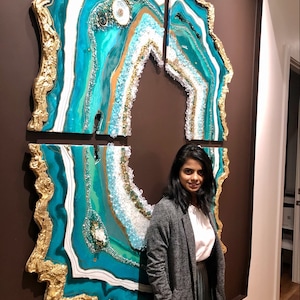 May include: A large, abstract wall art piece featuring a teal and gold agate design. The art is made up of four separate panels, each with a unique pattern. The art is displayed on a brown wall, with a person standing next to it.
