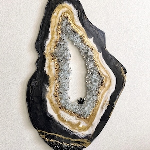 May include: An abstract wall art piece shaped like a geode, featuring concentric layers of black, white, and metallic gold resin. The center is filled with crushed clear glass fragments and small black crystal accents, creating a textured appearance.
