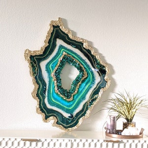 May include: A large green and gold resin art piece shaped like an agate geode. The geode has a white center with layers of green and gold resin around it. The geode is on a white shelf with a hexagonal pattern.