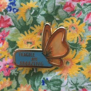 Puede incluir: Un broche de mariposa dorado con esmalte naranja y azul claro. El broche tiene las palabras "FRAGILE AND AMAZING" en letras doradas. El broche está sobre un fondo floral.