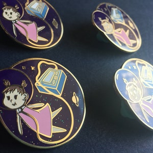 Puede incluir: Cuatro pines de esmalte de color dorado con temática espacial. Cada pin presenta a una niña de dibujos animados con un vestido rosa y un casco flotando en el espacio. El fondo es de color morado oscuro con purpurina dorada y una nave espacial azul y blanca.