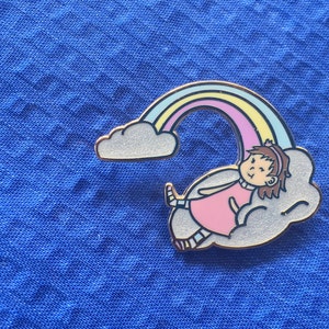 Puede incluir: Un pin de esmalte dorado con una niña de dibujos animados con cabello castaño acostada en una nube blanca con un arcoíris sobre ella. La niña lleva un vestido rosa y calcetines blancos.