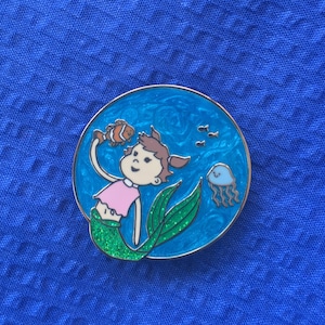 Puede incluir: Un pin de esmalte azul y verde con una sirena de dibujos animados de pelo castaño que sostiene un pez payaso. La sirena lleva una camiseta rosa y tiene una cola verde con purpurina. El fondo es un océano azul con una medusa y peces pequeños.
