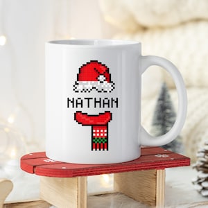 Könnte beinhalten: Weiße Keramik-Tasse mit pixeligem Weihnachtsdesign. Das Design zeigt einen roten Weihnachtsmannhut, einen roten Schal und den Namen "NATHAN" in Schwarz. Die Tasse steht auf einem kleinen roten Schlitten.