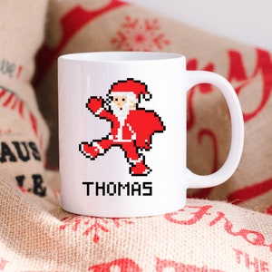 Könnte beinhalten: Weiße Keramiktasse mit einem pixelartigen Weihnachtsmann-Design und dem Namen "THOMAS" darunter gedruckt. Die Tasse steht auf einem Jutesack mit roter Beschriftung.
