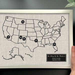 May include: A white framed map of the United States with black outlines of the states. The map is made of a light gray fabric and has black push pins with white text on them. The text on the pins includes "We Do", "First Kiss", "First Date", "We Met", and "First Home". The bottom right corner of the map has a black rectangle with white text that reads "The Story Of The Millan's, Matthew & Kelly, September 5, 2021."