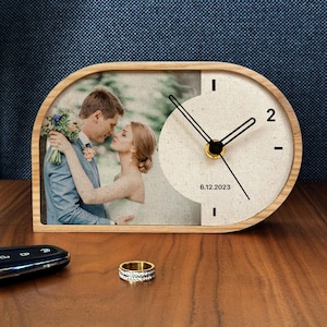 May include: Wooden desk clock with a photo of a couple and a clock face. The clock has black hands and the date 6.12.2023. A gold ring is in the foreground.