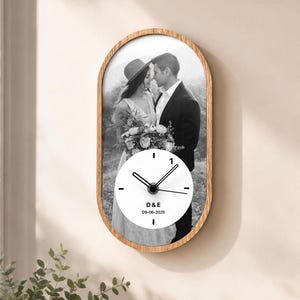 May include: An oval wall clock with a light wood frame featuring a black and white photograph of a couple. A circular white clock face is centered over the photo with black hands, the letters D & E, and the date 09-06-2025 printed in black.