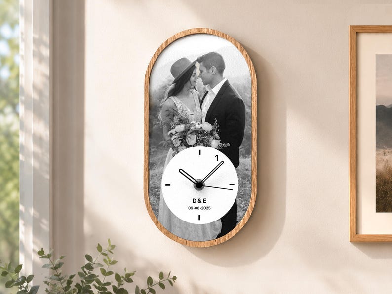 May include: An oval wall clock with a light wood frame featuring a black and white photograph of a couple. A circular white clock face is centered over the photo with black hands, the letters D & E, and the date 09-06-2025 printed in black.