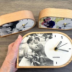 May include: Three semi-circular wooden desk clocks featuring custom black and white wedding photographs on the left side and a white clock face with black hands and the date 8.24.2024 printed on the right side of each clock face.