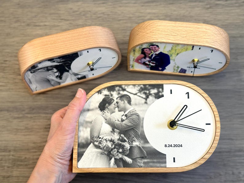 May include: Three semi-circular wooden desk clocks featuring custom black and white wedding photographs on the left side and a white clock face with black hands and the date 8.24.2024 printed on the right side of each clock face.