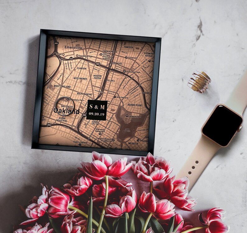 Puede incluir: Un mapa de Oakland, California, enmarcado en negro, con el texto "S & M 09.20.19" en un cuadro blanco. El mapa est&aacute; sobre una superficie blanca con un ramo de tulipanes rosas y un Apple Watch de oro rosa.