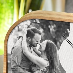 May include: A wooden wall clock featuring a black and white photograph of two people embracing. The clock face is integrated into the image, showing thin black hands against the monochrome portrait of the couple in a close-up, affectionate pose.