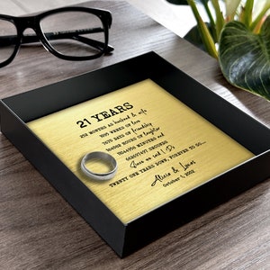 May include: A gold-colored plaque with black text commemorating 21 years of marriage. The plaque is in a black frame and includes the text "21 Years", "252 months as husband & wife", "1095 weeks of love", "7670 days of friendship", "184082 hours of laughter", "11044956 minutes", "662697471 seconds", "Since we said I Do", "Twenty one years down, forever to go...", "Alicia & Lucas", "October 1, 2002". A silver ring is placed on the plaque.