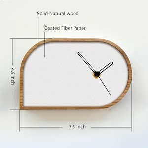 May include: A wall clock with a solid natural wood frame and a white coated fiber paper face. The clock has black hands and measures 7.5 inches in width by 4.9 inches in height. The design features a rounded, asymmetrical shape.