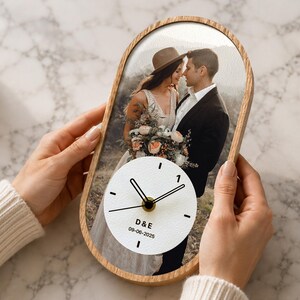 May include: An oval-shaped wall clock with a light wood frame featuring a printed wedding photograph of a couple. The clock face is white with black hands and markers, displaying the initials D&E and the date 09-06-2025 in black text.