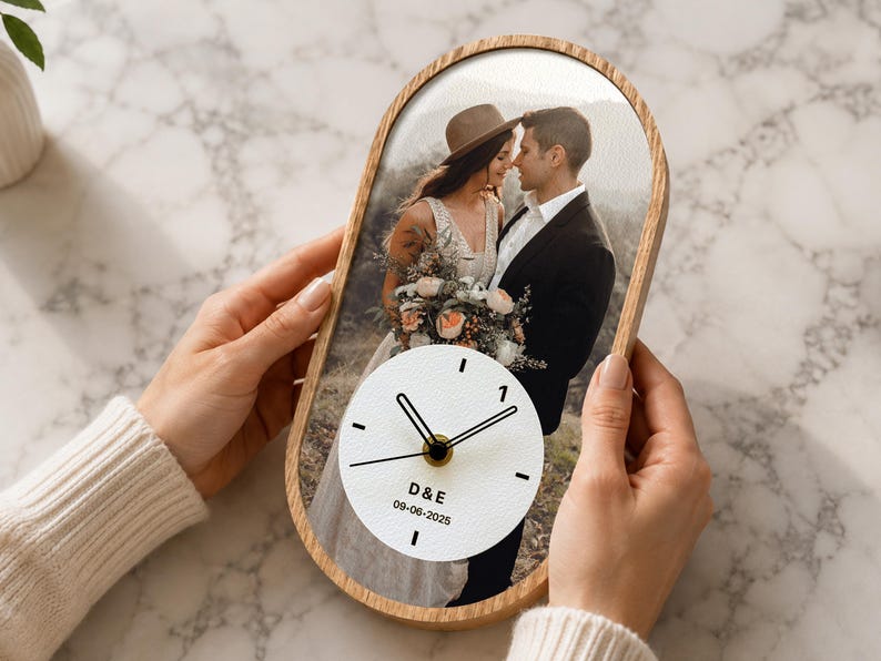 May include: An oval-shaped wall clock with a light wood frame featuring a printed wedding photograph of a couple. The clock face is white with black hands and markers, displaying the initials D&E and the date 09-06-2025 in black text.