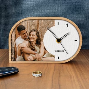 May include: A semi-oval wooden desk clock featuring a custom photograph of a couple embracing in a forest setting. The white clock face displays black hour markers, black hands, and the printed date 12.14.2023 on the right side of the frame.