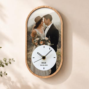 May include: An oval wall clock with a wooden frame featuring a printed photograph of a couple in wedding attire. The clock face is white with black hands and markers, displaying the text D&E and the date 09-06-2025 in black lettering.