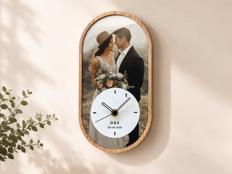 May include: An oval wall clock with a wooden frame featuring a printed photograph of a couple in wedding attire. The clock face is white with black hands and markers, displaying the text D&E and the date 09-06-2025 in black lettering.