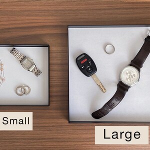 Puede incluir: Dos bandejas, una etiquetada como "Peque&ntilde;a" y otra como "Grande", con varios art&iacute;culos en su interior. La bandeja peque&ntilde;a contiene un reloj plateado, un collar de perlas y dos anillos plateados. La bandeja grande contiene un reloj de pulsera de cuero marr&oacute;n, una llave de coche y un anillo plateado.
