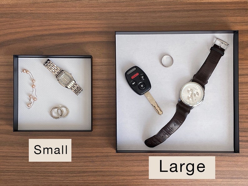 Puede incluir: Dos bandejas, una etiquetada como "Peque&ntilde;a" y otra como "Grande", con varios art&iacute;culos en su interior. La bandeja peque&ntilde;a contiene un reloj plateado, un collar de perlas y dos anillos plateados. La bandeja grande contiene un reloj de pulsera de cuero marr&oacute;n, una llave de coche y un anillo plateado.