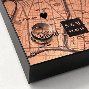 May include: A black shadow box with a map of Oakland, California. The map is printed on a copper-colored background. Two silver wedding bands are placed on the map. The date "09.20.19" and the initials "S&M" are printed on the map.