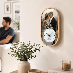 May include: An oval-shaped wall clock with a light wood frame featuring a printed photograph of a couple. The clock face is white with black hands and includes the text D&E and 30-08-2020. The design combines a personalized photo with a functional timepiece.