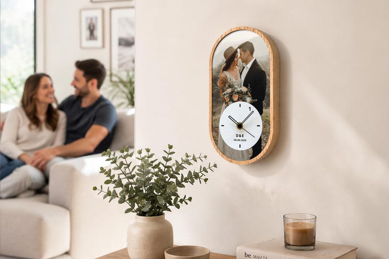 May include: An oval-shaped wall clock with a light wood frame featuring a printed photograph of a couple. The clock face is white with black hands and includes the text D&E and 30-08-2020. The design combines a personalized photo with a functional timepiece.