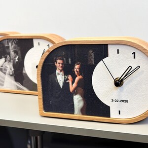 May include: A horizontal, semi-oval wooden desk clock featuring a custom black and white wedding photograph on the left side and a white clock face on the right. The clock has black hands, the number 1, and the printed date 2-22-2025.
