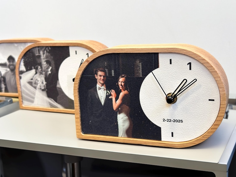 May include: A horizontal, semi-oval wooden desk clock featuring a custom black and white wedding photograph on the left side and a white clock face on the right. The clock has black hands, the number 1, and the printed date 2-22-2025.