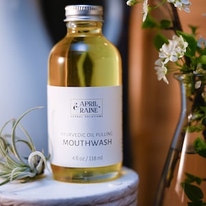 May include: A clear glass bottle with a silver lid and a white label. The label reads "APRIL & RAINE HERBAL SOLUTIONS AYURVEDIC OIL PULLING MOUTHWASH 4 fl oz/118 ml".