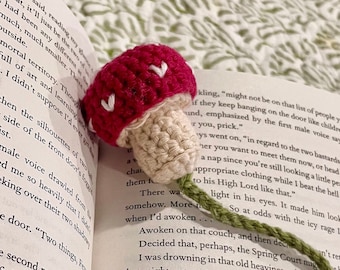 May include: A crocheted mushroom bookmark with a red cap and white spots, a green stem, and a green leaf. The bookmark is placed on an open book with text about a fantasy world.