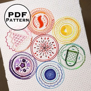May include: A set of seven colourful embroidery patterns on white fabric. Each pattern is a circle with a different design and colour scheme. The patterns are arranged in a circle around the centre of the fabric.