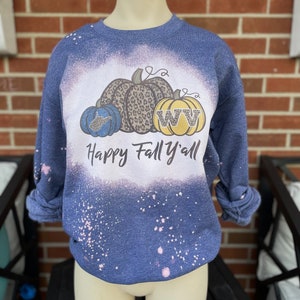 May include: A blue sweatshirt with a white and pink splattered design. The front features three pumpkins with the text "Happy Fall Y'all". The pumpkins have leopard print and state emblems.
