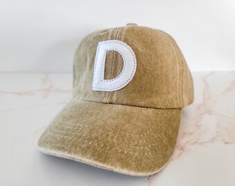 May include: A khaki baseball cap featuring a white embroidered letter "D". The cap has a curved peak and an adjustable strap. The hat is made of a soft, worn material.