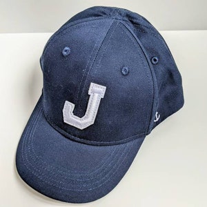 May include: A navy blue baseball cap with a white felt letter "J" on the front and a small white anchor on the side.