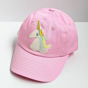 May include: Pink baseball cap with a unicorn appliqué. The unicorn is white with a gold horn and rainbow mane.
