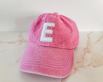 May include: A pink baseball cap featuring a white embroidered letter "E". The cap has a curved peak and an adjustable strap. The hat is made of a soft, faded fabric.