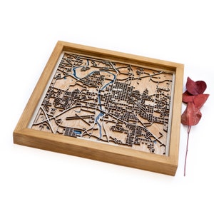 May include: A wooden map of a city in a light wood frame. The map is laser cut and shows the streets and waterways of the city. The map is a 3D design and has a blue water feature.